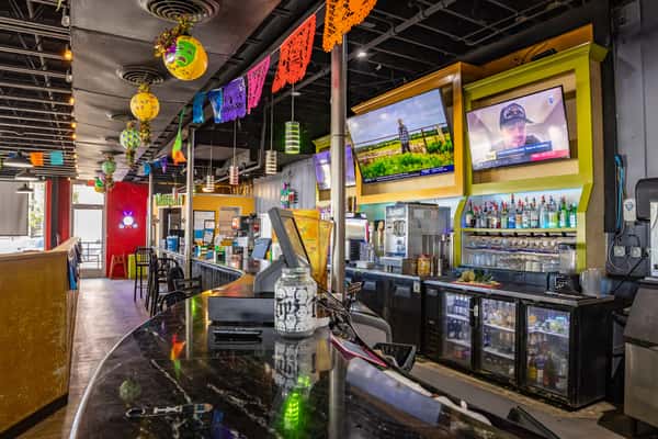Colorful Mexican restaurant bar interior with papel picado streamers, hanging lanterns, flat-screen TVs, and a fully stocked liquor display — featuring vibrant decor, festive ambiance, and modern casual dining vibe.
