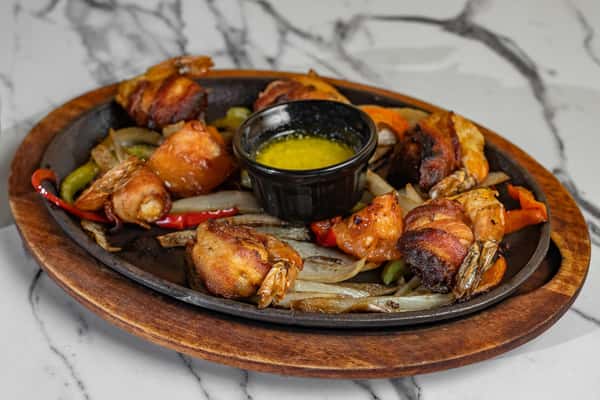 Sizzling platter of bacon-wrapped shrimp served over grilled peppers and onions with a side of melted butter sauce.