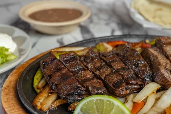 Sizzling steak fajitas served on a hot skillet with grilled onions and peppers, accompanied by lime wedges, refried beans, sour cream, and tortillas — a bold Tex-Mex classic.
