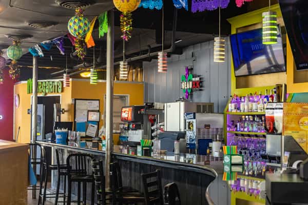 Lively Mexican bar with hanging decorations, papel picado streamers, vibrant lighting, and a stocked liquor display — festive setting for cocktails and casual nightlife.