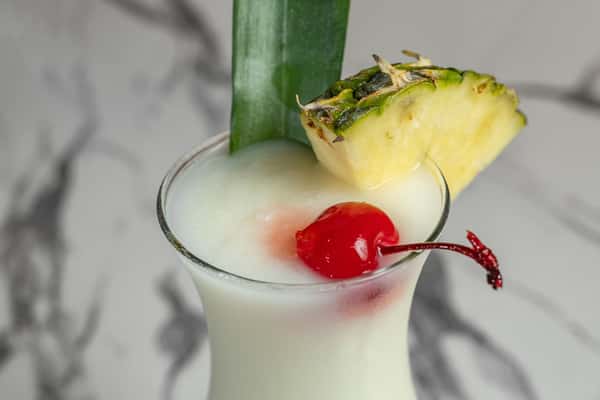 Creamy piña colada served in a curved cocktail glass, garnished with a pineapple wedge, maraschino cherry, and tropical leaf — refreshing island-style drink perfect for summer menus or tropical-themed cocktails.