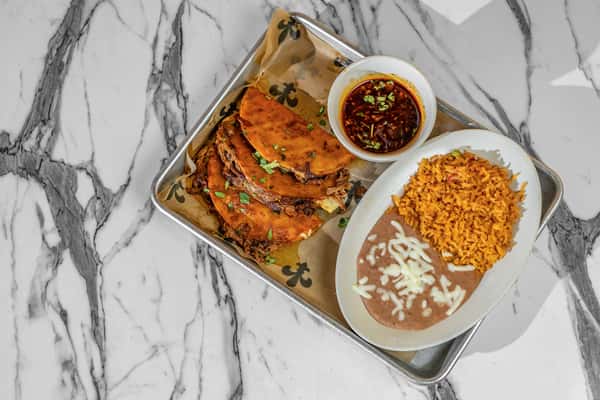 Plated birria tacos with crispy, golden shells filled with slow-cooked beef, served with rich consommé for dipping, alongside refried beans with melted cheese and seasoned Mexican rice — traditional Jalisco-style street food presented on a modern tray.