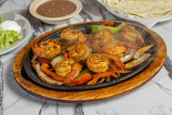 Sizzling shrimp fajitas served on a hot cast iron skillet with grilled onions and bell peppers, accompanied by refried beans, tortillas, and fresh lettuce.