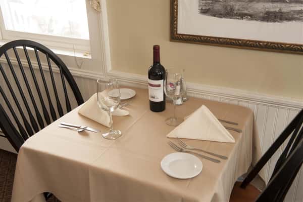 Empty dining room with tables covered in beige tableclothes surrounded by wooden brown and black chairs with wine glasses and a bottle of wine on top