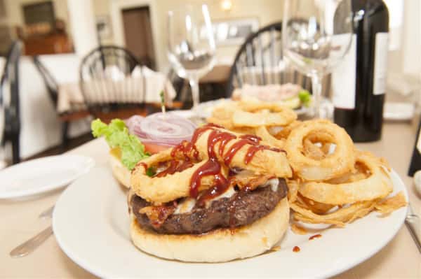 Burger covered with cheese, bacon, and onion rings drizzled with red sauce on white plate next to wine glasses