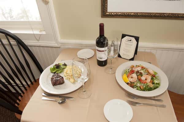 Two dishes with a steak and a shrimp salad on a beige table next to two wine glasses