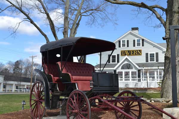 Exterior of White paneled building with "Tavern" sign in middle next to old wooden carriage