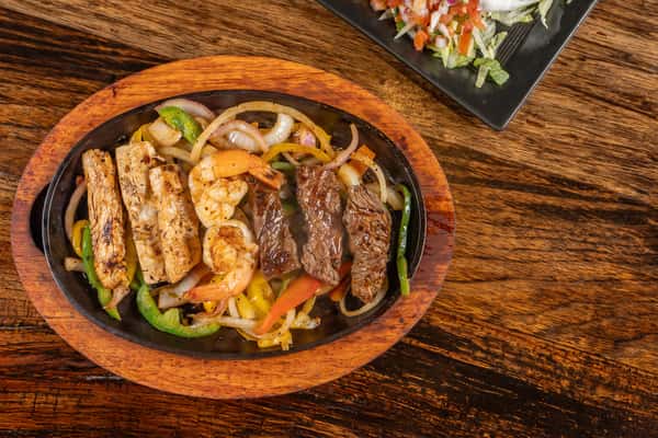 Sizzling fajita platter with grilled chicken, shrimp, and steak served on a bed of sautéed onions and bell peppers in a hot cast iron skillet.
