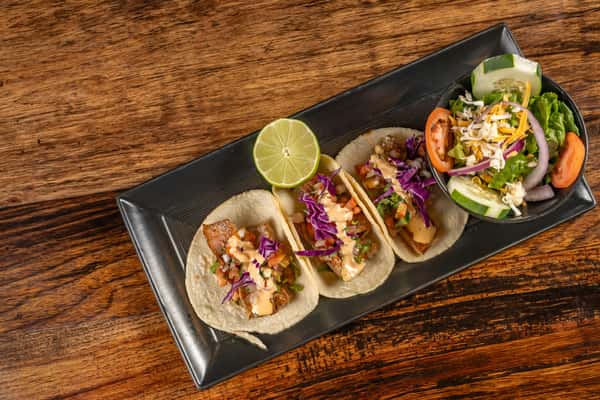 Three soft corn tortillas filled with grilled fish, shredded cabbage, pico de gallo, and creamy chipotle sauce, served with a lime wedge and a fresh garden salad with cucumber, tomato, and shredded cheese.