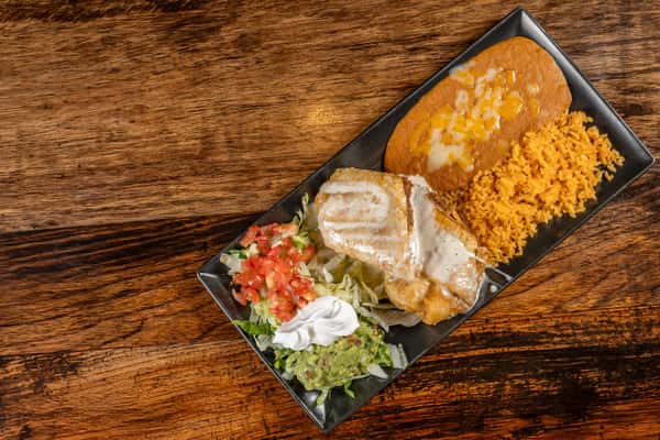Smothered chicken burrito topped with queso sauce, served with refried beans, Mexican rice, shredded lettuce, guacamole, sour cream, and pico de gallo on a rectangular plate.