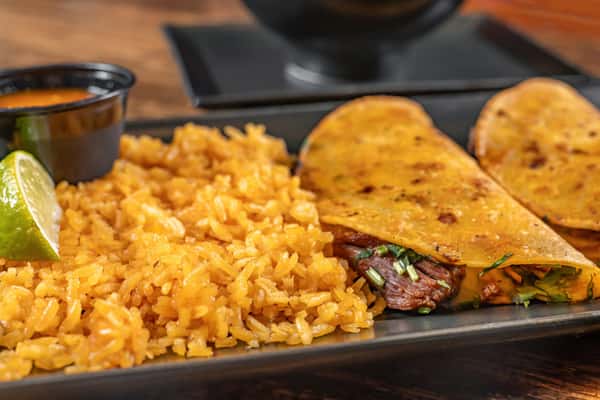 Golden birria tacos filled with shredded beef and cheese, served with Mexican rice, a lime wedge, spicy salsa, and rich consommé for dipping.