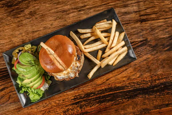 Grilled burger with melted cheese on a toasted brioche bun, served with French fries, sliced avocado, jalapeños, and fresh vegetables on a black rectangular plate.