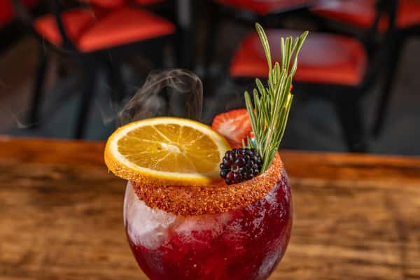 Vibrant red craft cocktail in a round glass with a chili-salt rim, garnished with a fresh orange slice, blackberry, strawberry, rosemary sprig, and a hint of smoke for a dramatic presentation.