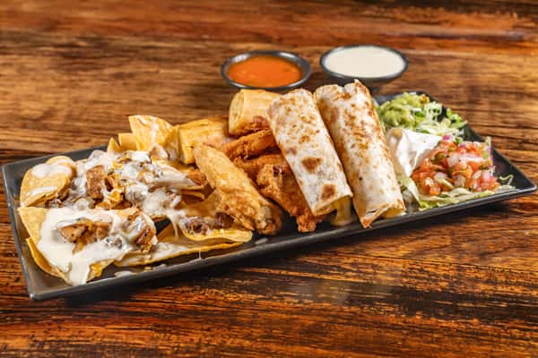 Loaded nachos with melted cheese and grilled chicken, crispy taquitos, fried chicken tenders, and mini burritos, served with guacamole, sour cream, pico de gallo, and dipping sauces on a rectangular black plate.