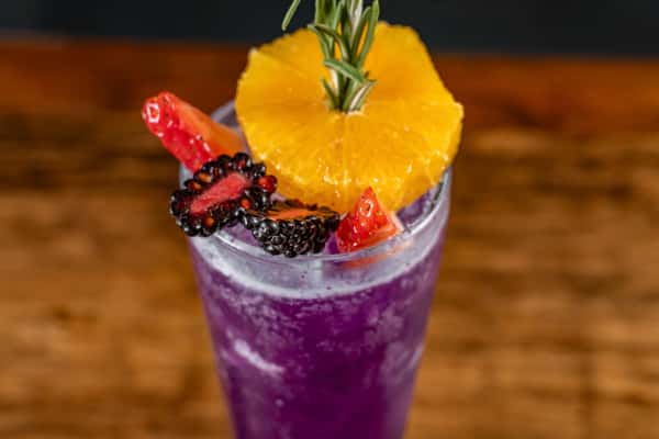 Vibrant purple cocktail served in a tall glass, garnished with fresh orange slice, blackberries, strawberries, and a sprig of rosemary, over ice—perfect for a refreshing and colorful signature drink.