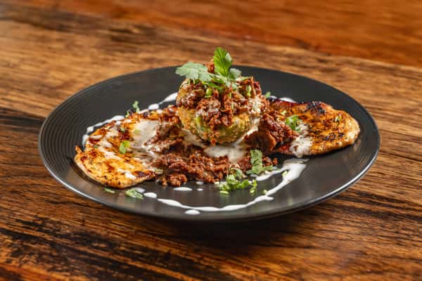Grilled stuffed avocado topped with seasoned ground beef and fresh cilantro, served with grilled zucchini and creamy drizzle on a black plate.