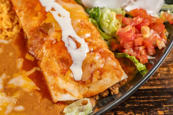 Cheesy beef enchilada smothered in red sauce and drizzled with sour cream, served with refried beans, Mexican rice, shredded lettuce, and pico de gallo on a round plate.