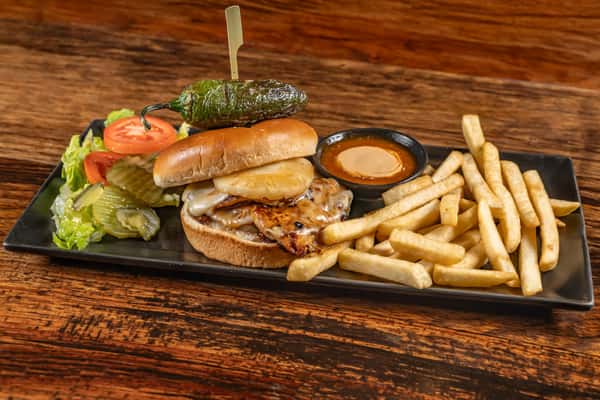 Grilled chicken sandwich with melted cheese, pineapple slice, and roasted jalapeño on a toasted bun, served with crispy French fries, dipping sauce, and a fresh side salad of lettuce, tomato, and pickles on a black rectangular plate.