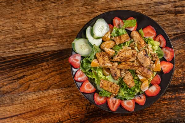 Fresh grilled chicken salad with mixed greens, mandarin orange slices, sliced strawberries, cucumber rounds, and chopped walnuts, served on a black plate over rustic wood—perfect for a light and vibrant lunch option.