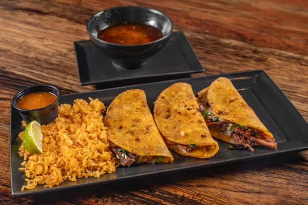 Three crispy birria tacos filled with shredded beef and melted cheese, served with Mexican rice, spicy dipping consommé, lime wedge, and red salsa on a rectangular plate.