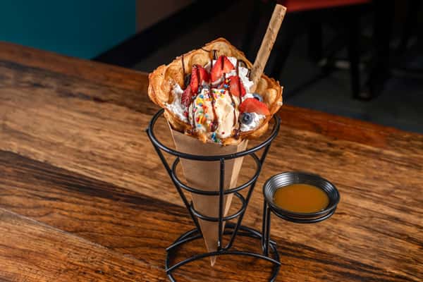 Golden waffle cone filled with vanilla ice cream, fresh strawberries and blueberries, whipped cream, rainbow sprinkles, chocolate drizzle, and a cookie stick, served with caramel sauce on the side.