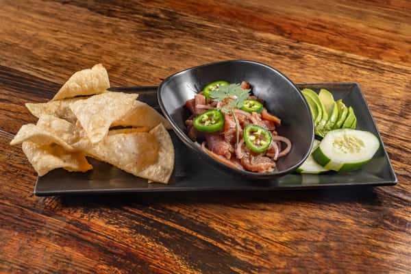Spicy tuna ceviche garnished with jalapeños and cilantro, accompanied by crispy tortilla chips, avocado, and cucumber slices on a black serving plate.