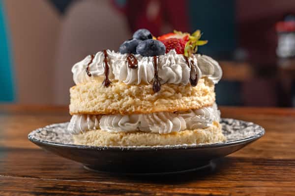 Moist layered vanilla cake filled and topped with whipped cream, drizzled with chocolate sauce, and garnished with fresh blueberries and a strawberry on a decorative plate—perfect as a rich and indulgent dessert option.
