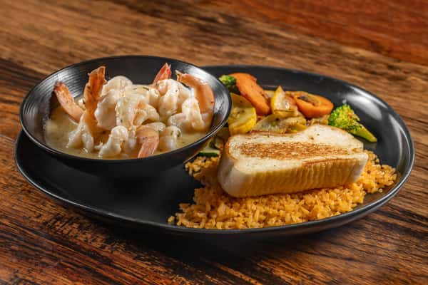 Creamy garlic shrimp in a bowl, accompanied by seasoned rice, grilled vegetables, and a slice of toasted bread on a black dinner plate.