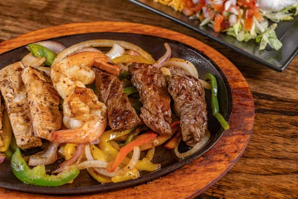 Sizzling trio fajitas with grilled shrimp, steak, and chicken, served over sautéed onions and bell peppers on a hot cast iron skillet with a wooden base.