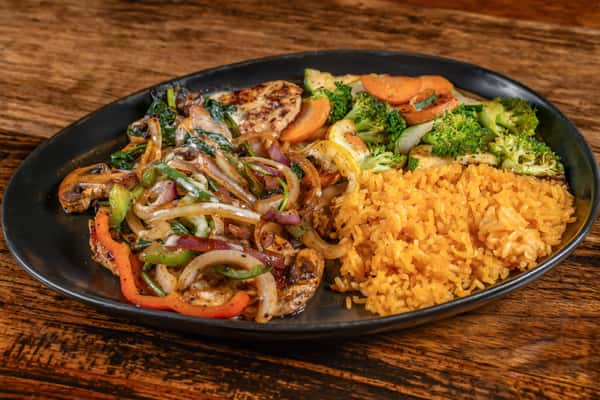 Grilled chicken breast topped with sautéed onions and bell peppers, served with seasoned Mexican rice and a colorful mix of steamed vegetables on a black oval plate.