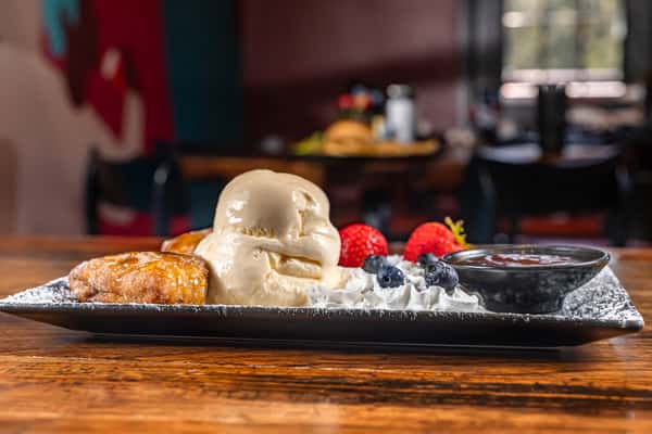 Vanilla ice cream served with cinnamon-sugar pastry, fresh strawberries, blueberries, whipped cream, and a side of chocolate sauce on a rustic black platter.
