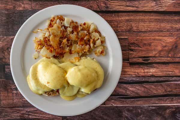Classic eggs Benedict topped with creamy hollandaise sauce, served alongside a portion of golden-brown home fries on a white plate — a popular brunch favorite perfect for cozy breakfast restaurants.