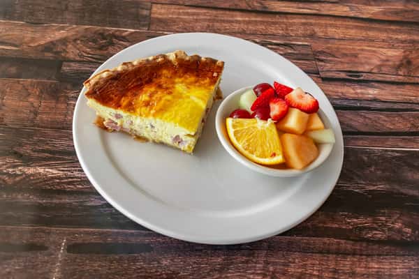 Slice of ham and cheese quiche with a golden crust, served with a colorful fruit cup featuring orange, strawberries, melon, and grapes — a comforting and balanced brunch entrée.