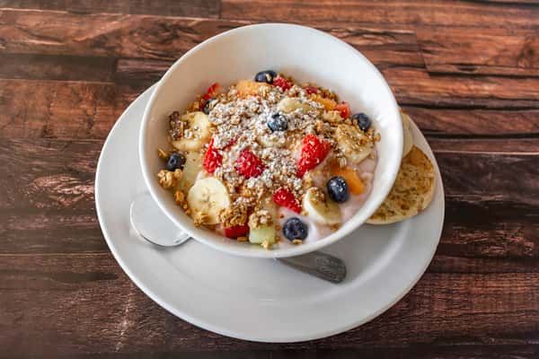 Colorful fruit and granola parfait made with fresh bananas, strawberries, blueberries, and cantaloupe over creamy yogurt, served with toasted English muffin halves — a vibrant and healthy breakfast option.
