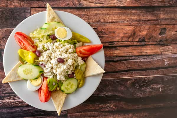 Fresh Greek salad topped with crumbled feta, Kalamata olives, cucumber, tomato wedges, pepperoncini, and hard-boiled egg slices, served with warm pita triangles — a vibrant Mediterranean-style lunch option.