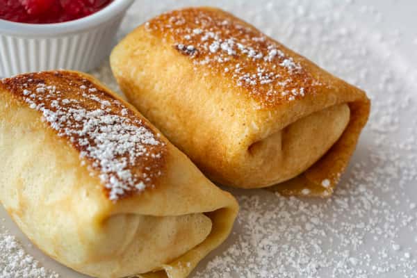 Close-up of two golden, pan-seared cheese blintzes dusted with powdered sugar, served with a side of sweet strawberry compote — a rich and creamy Eastern European breakfast or brunch dessert.