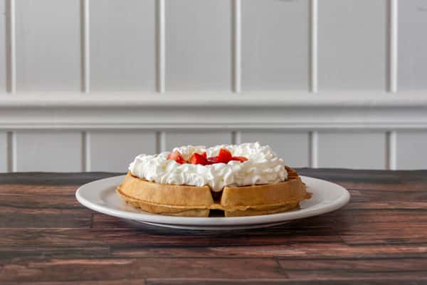 Fluffy golden waffle topped with a generous swirl of whipped cream and fresh strawberry slices, served on a white plate — a sweet and indulgent breakfast or brunch treat.