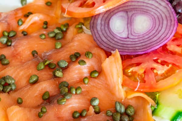 Close-up of silky smoked salmon (lox) topped with capers and served with sliced tomatoes, red onions, and cucumbers — a flavorful and fresh brunch favorite with Mediterranean flair.