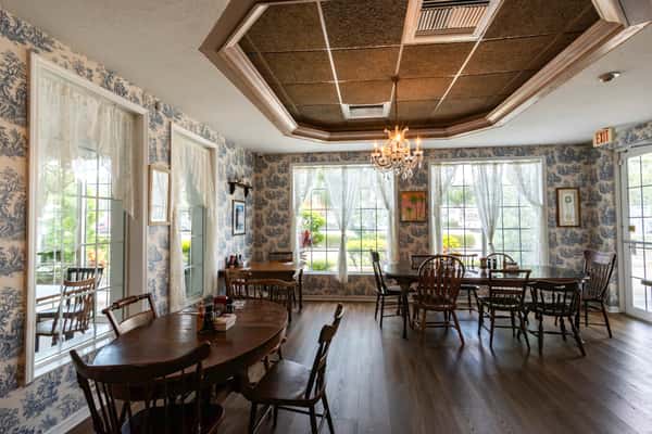 Charming dining area with vintage-style wallpaper, natural lighting from large windows, wooden tables and chairs, and an elegant chandelier — cozy ambiance ideal for a relaxing breakfast or lunch experience.