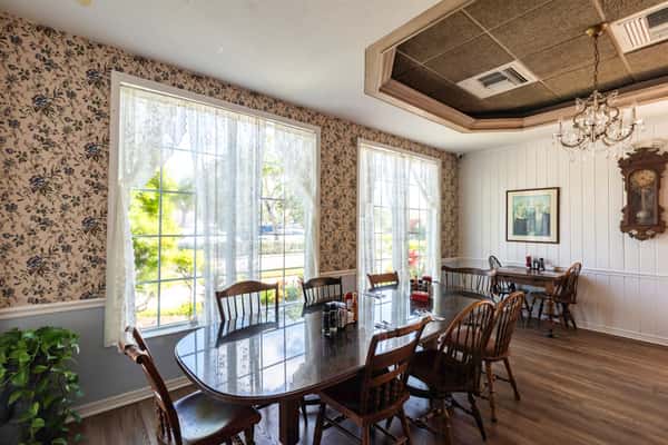 Elegant dining space with floral wallpaper, lace curtains, wooden furniture, and a chandelier — a vintage-inspired ambiance ideal for relaxed brunch or lunch settings.