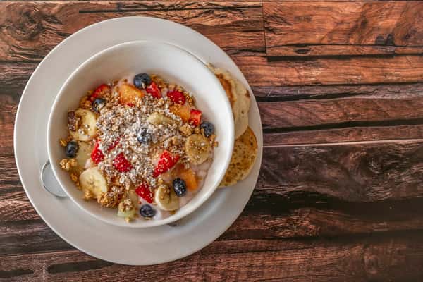 Creamy yogurt parfait topped with fresh bananas, strawberries, blueberries, and crunchy granola, lightly dusted with powdered sugar, served with toasted English muffin halves — a wholesome and colorful breakfast choice.