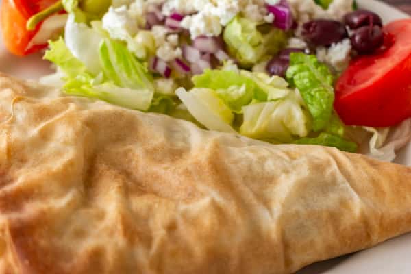 Crispy baked spanakopita triangle next to a fresh Greek salad with feta cheese, olives, lettuce, tomato, and red onion — a Mediterranean-inspired vegetarian plate.