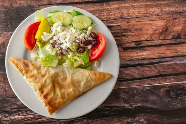 Crispy spanakopita triangle served with a Greek side salad featuring crumbled feta, Kalamata olives, cucumber slices, pepperoncini, and fresh tomato wedges — a Mediterranean-inspired vegetarian dish perfect for brunch or lunch menus.