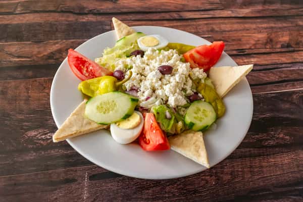 Classic Greek salad with crumbled feta cheese, Kalamata olives, tomatoes, cucumbers, pepperoncini, and hard-boiled egg slices, served with pita wedges on a white plate — a fresh and Mediterranean-inspired lunch or appetizer.