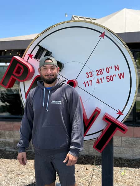 A man wearing a backward cap and a dark hoodie stands in front of a large circular "The POINT" sign displaying geographic coordinates outside a building on a sunny day.