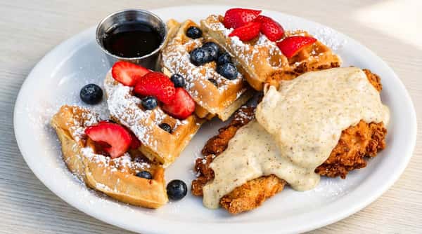 Waffles with strawberries, blueberries, powdered sugar, and a side of fried chicken with gravy, and maple syrup.