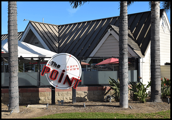 Exterior view of a restaurant building with palm trees and a large "The POINT" sign displaying coordinates in front.