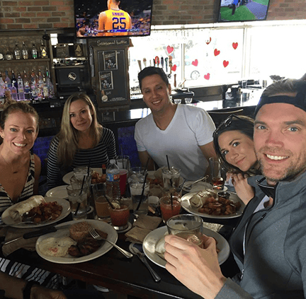 Group of five friends smiling at a restaurant table filled with brunch plates and drinks, with a TV and bar area in the background.