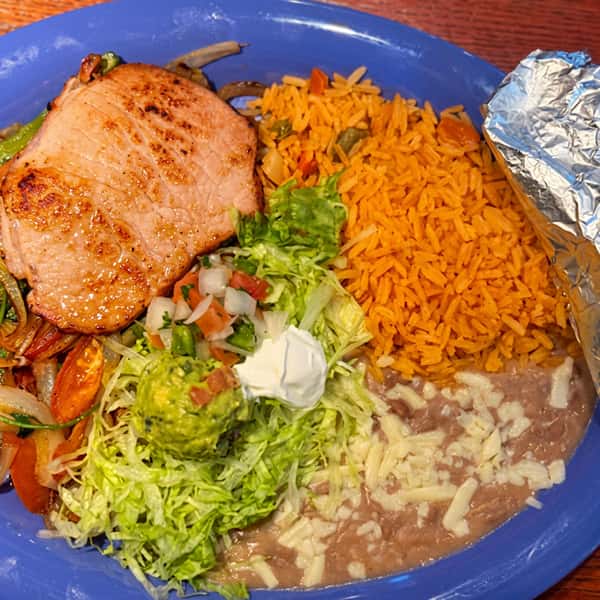Grilled pork chop served with Mexican rice, refried beans topped with melted cheese, grilled vegetables, fresh salad, guacamole, sour cream, and pico de gallo at El Cazo Mexican Restaurant.