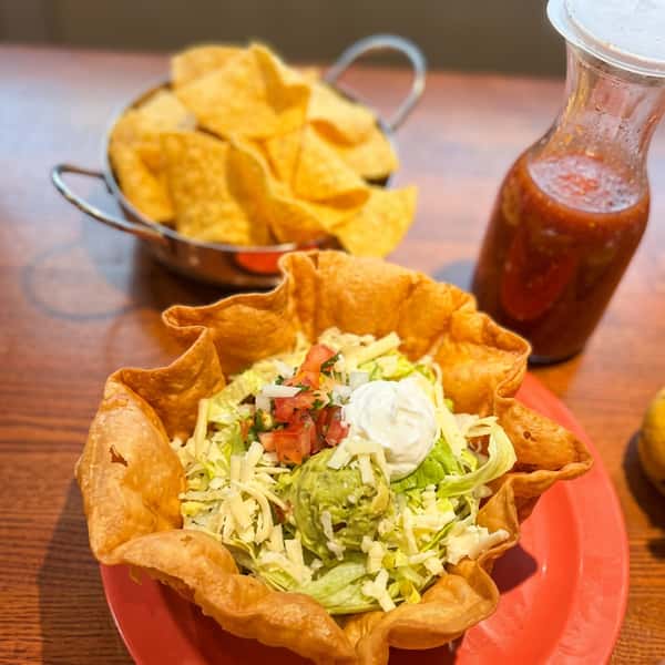 Crispy taco salad served in a golden tortilla shell bowl with fresh lettuce, shredded cheese, guacamole, sour cream, and pico de gallo, accompanied by tortilla chips and house-made salsa at El Cazo Mexican Restaurant.